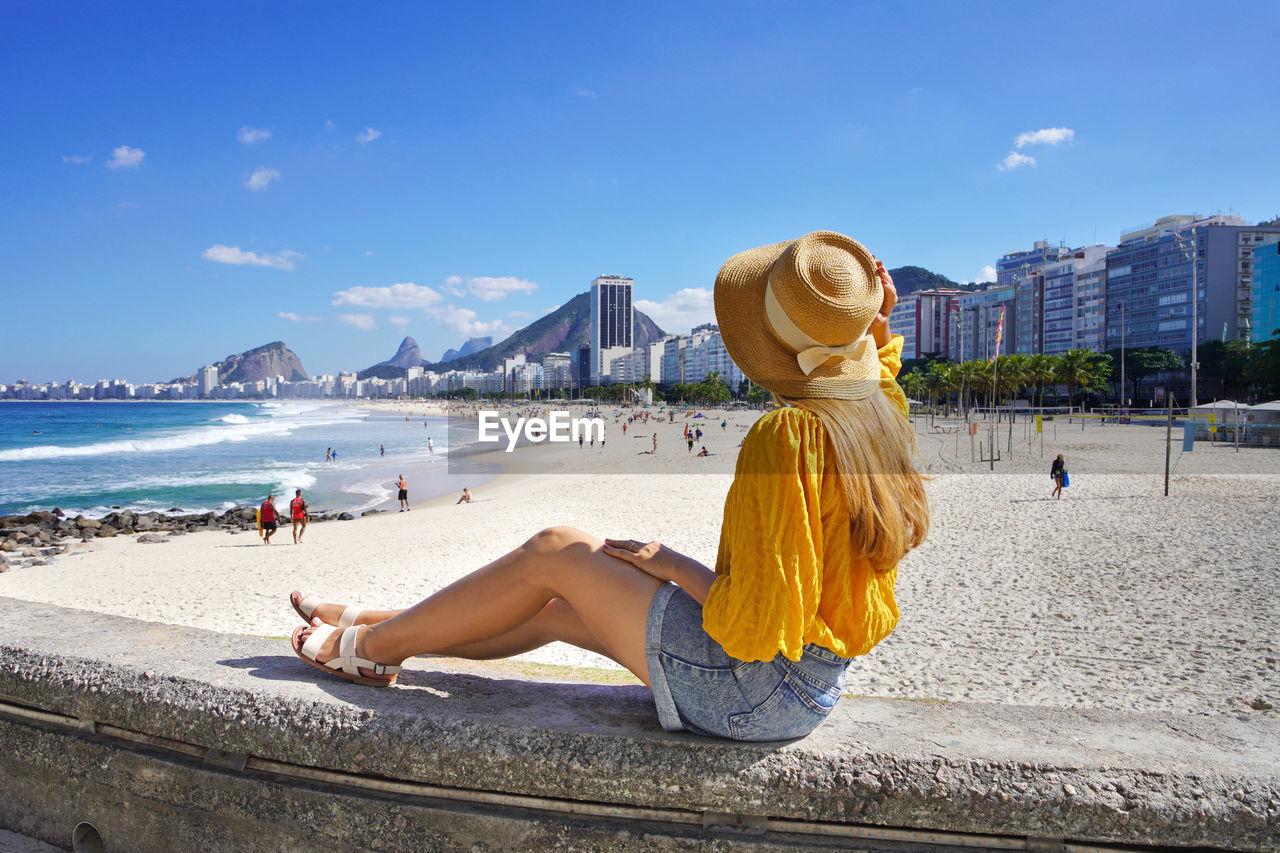 Beautiful fashion girl sitting on wall enjoying view of copacabana beach. summer vacation in brazil.