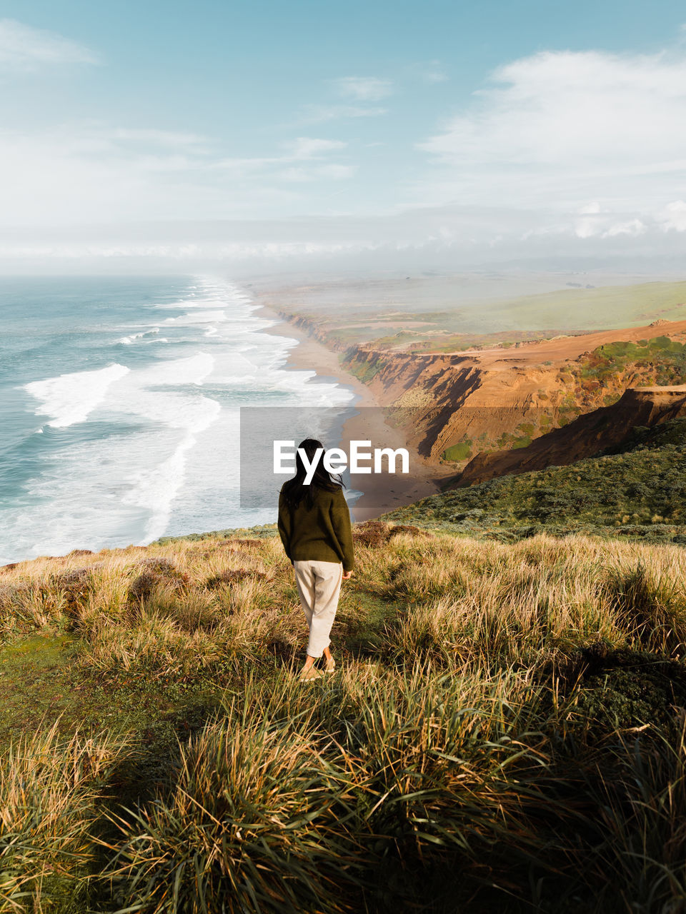 Back view of woman standing in solitude on high cliff meadow of point reyes national seashore observing majestic view of ocean in california