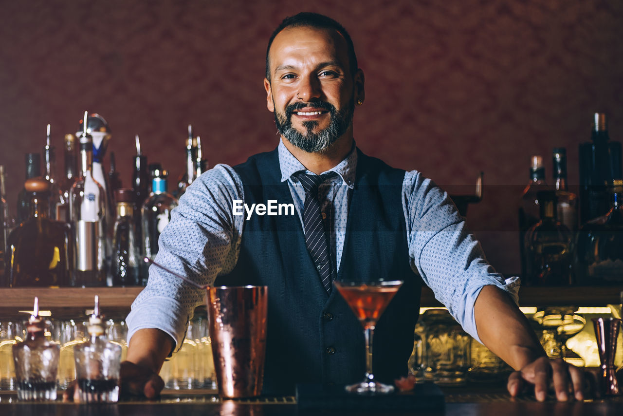 Portrait of smiling mature bartender standing by cocktail on bar counter