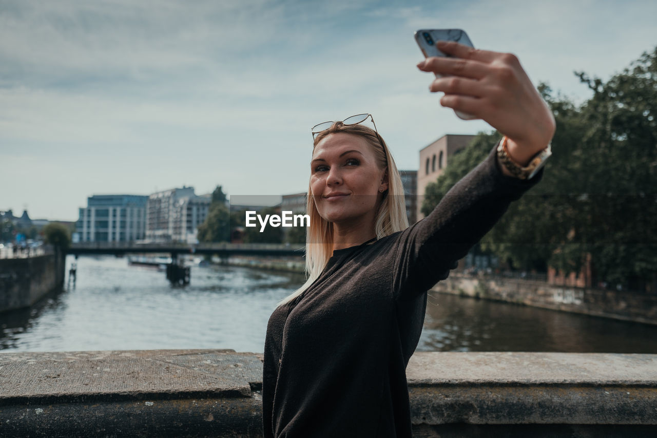 Smiling young woman taking selfie while standing in city