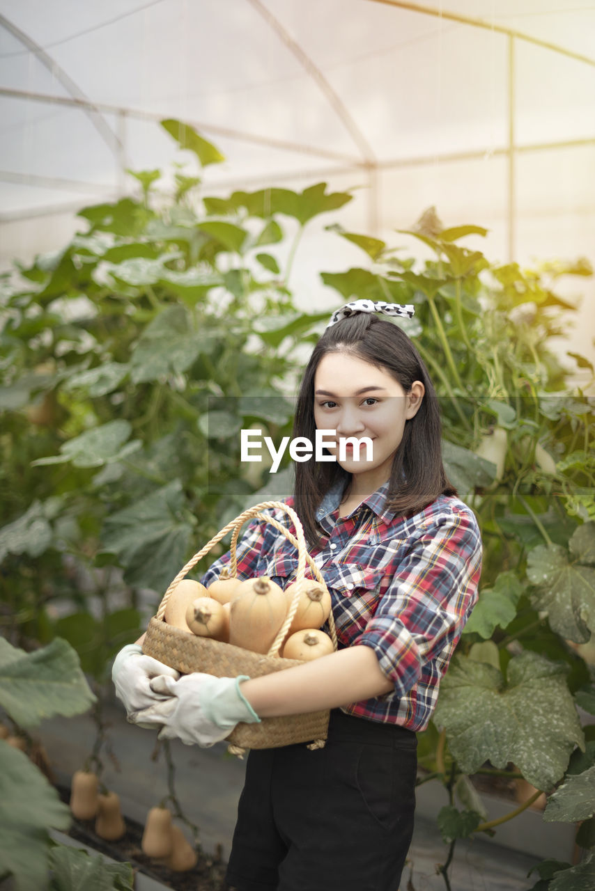 Portrait of smiling young woman carrying vegetables while standing against plants in greenhouse