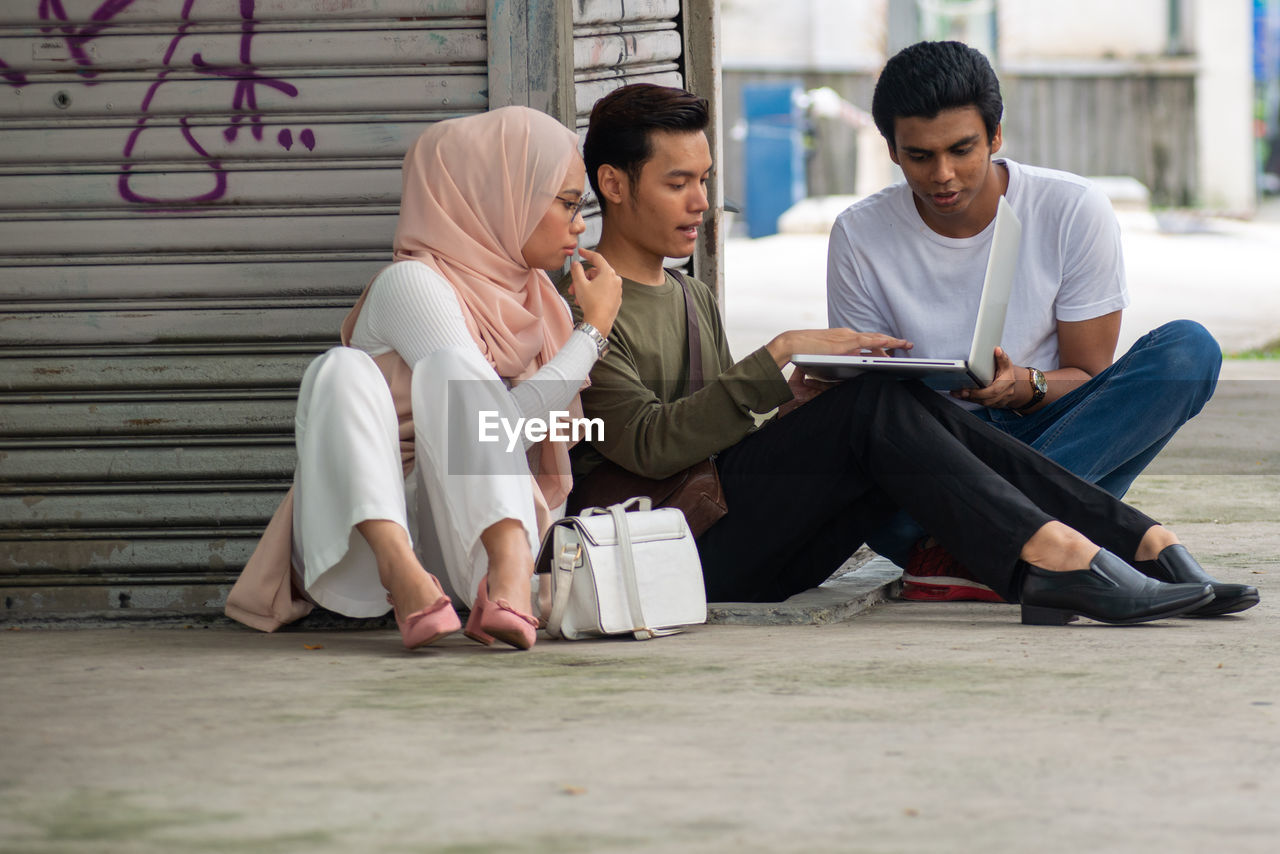 YOUNG MAN USING MOBILE PHONE WHILE SITTING ON LAPTOP OUTDOORS