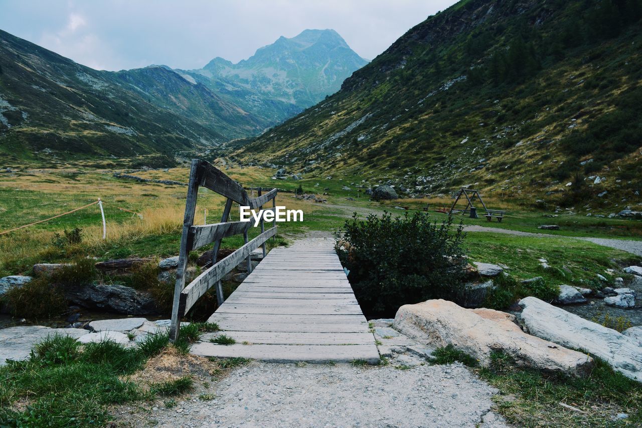 Wooden walkway on field by mountains against sky