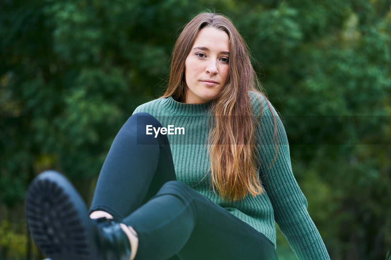 Portrait of a young caucasian woman with brown hair in a park with the green of the trees in the background dressed in a green sweater, dark pants and military boots