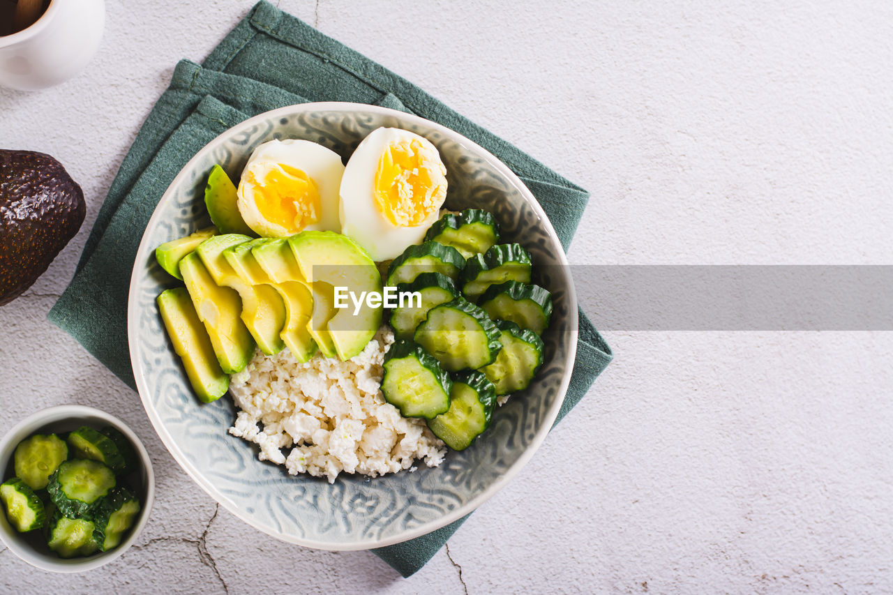 Bowl with cottage cheese, cucumber and avocado slices and boiled egg on the table top view