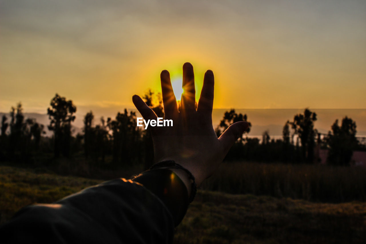 Cropped hand covering sun in sky during sunset