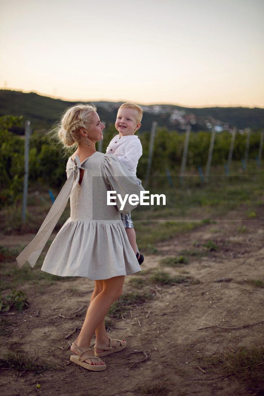 Blonde woman in a dress holds her baby boy in her arms in a vineyard during sunset