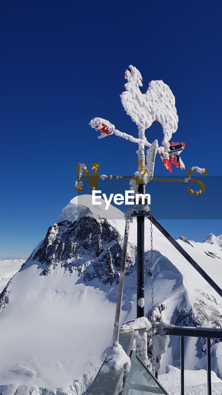 Low angle view of frozen weather vane against mountains and clear blue sky during winter