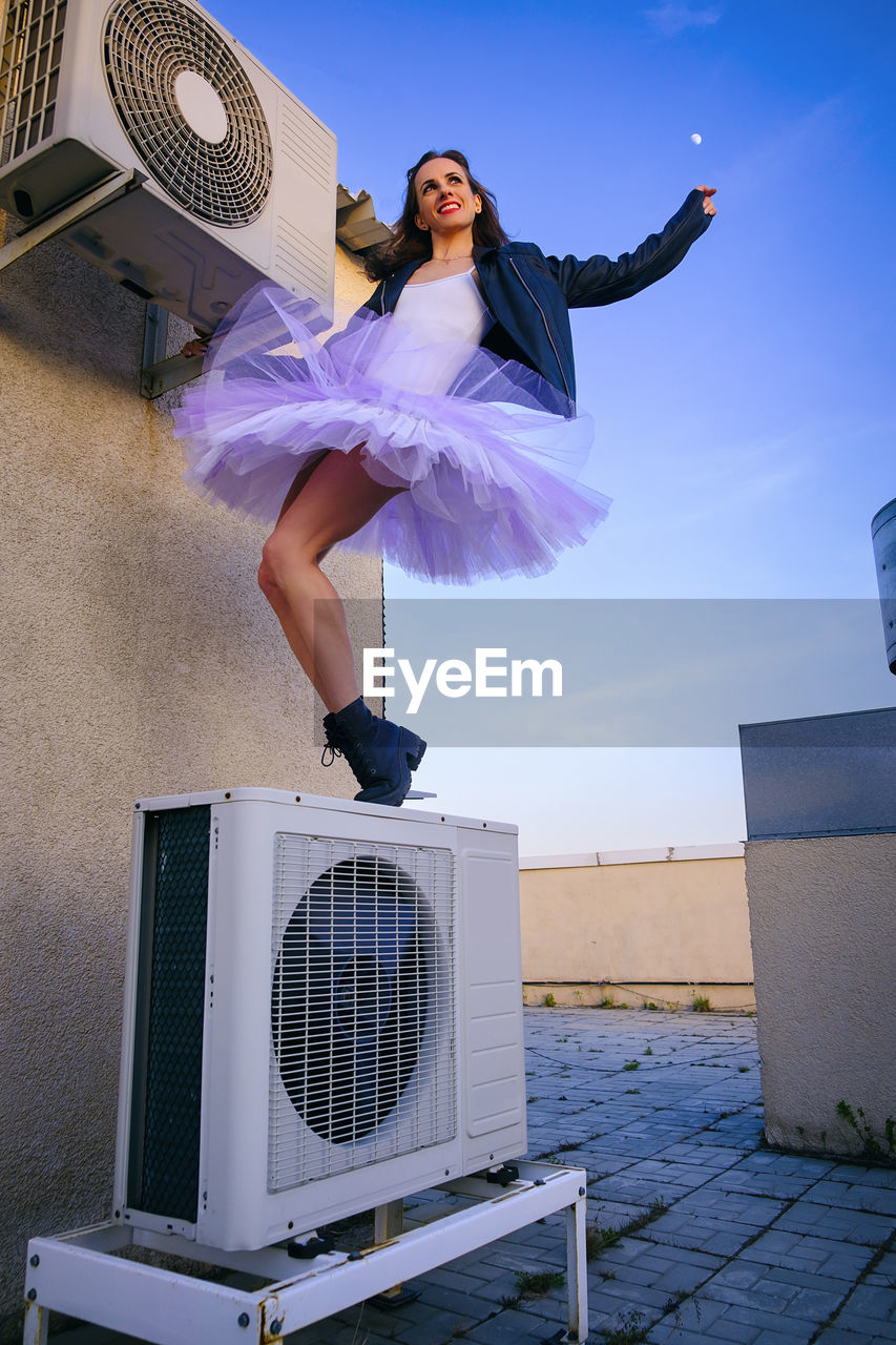 A ballerina in tutu, jacket and boots climbed on the air conditioner and poses against the sky