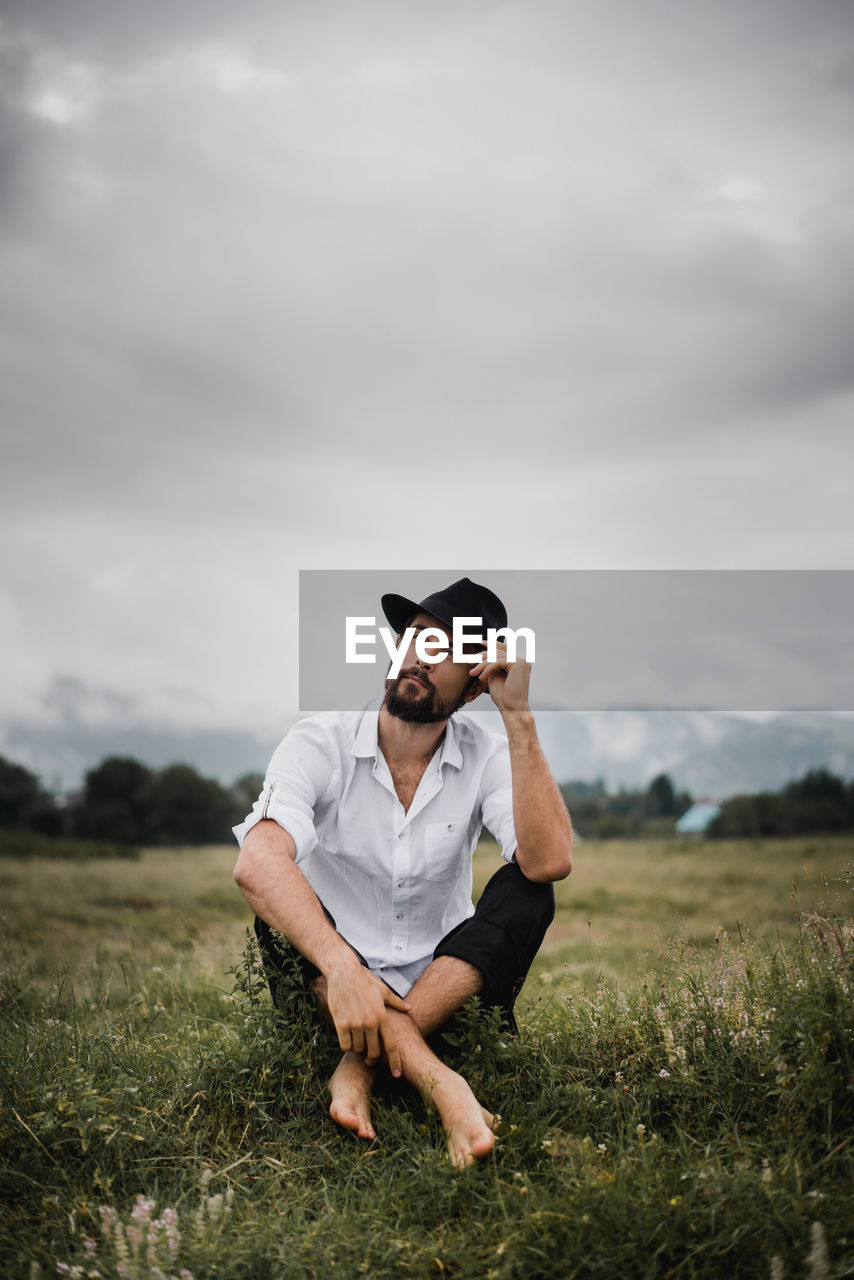 Thoughtful young man sitting on grassy field against cloudy sky