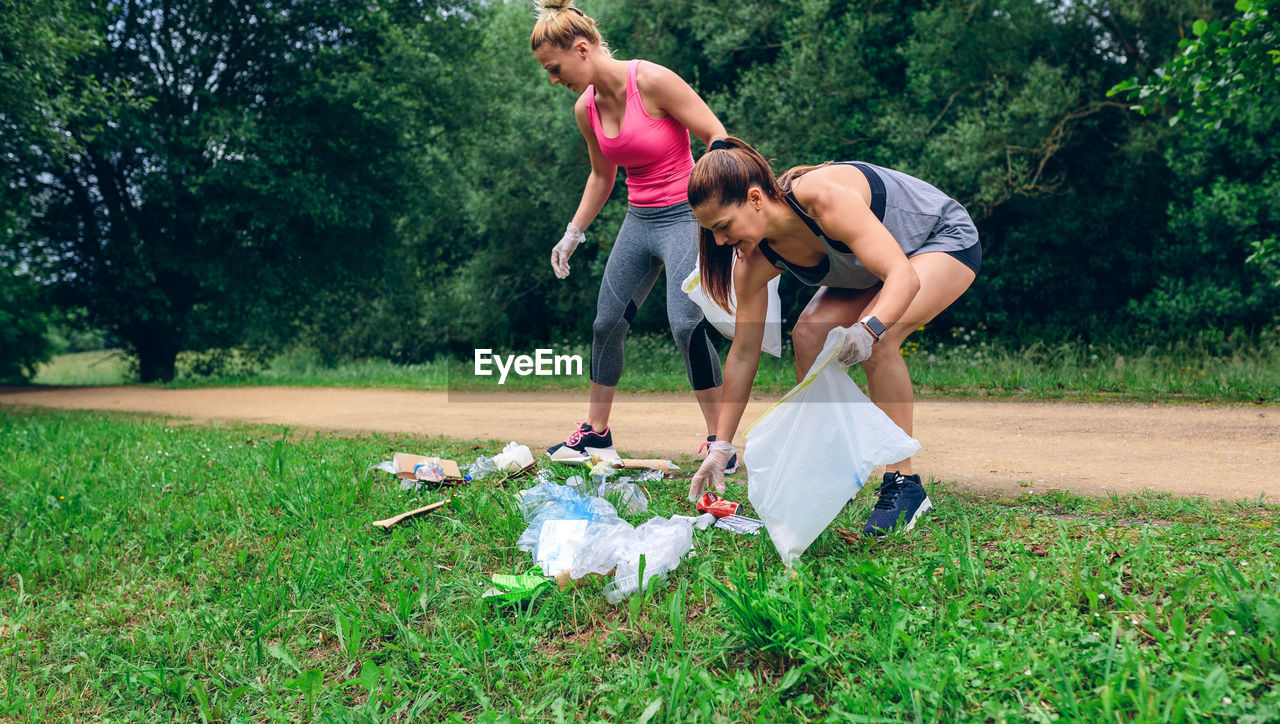 Full length of happy women picking up garbage while crouching on grass against trees