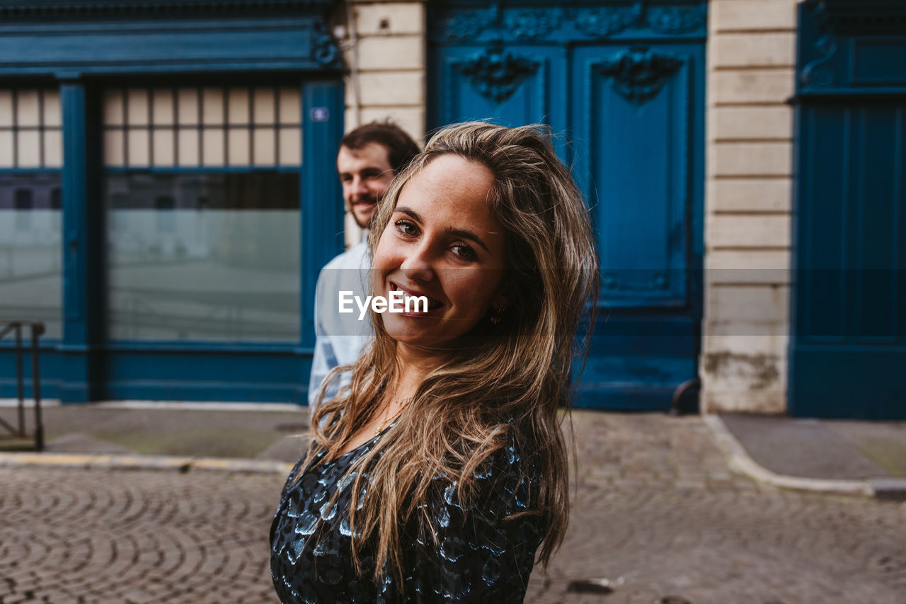 Happy young woman looking at camera in stylish outfit followed by smiling boyfriend walking on city street with old building in background during romantic holidays in france