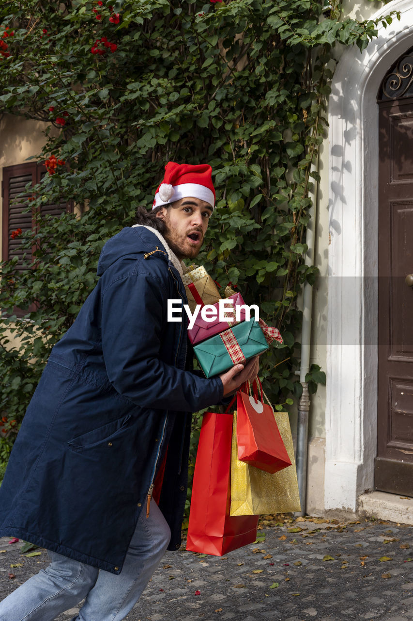 Young man with santa claus hat has a surprised expression and brings his newly purchased gifts.