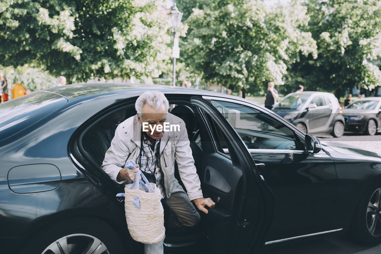 Senior man holding bag getting out from black car in city