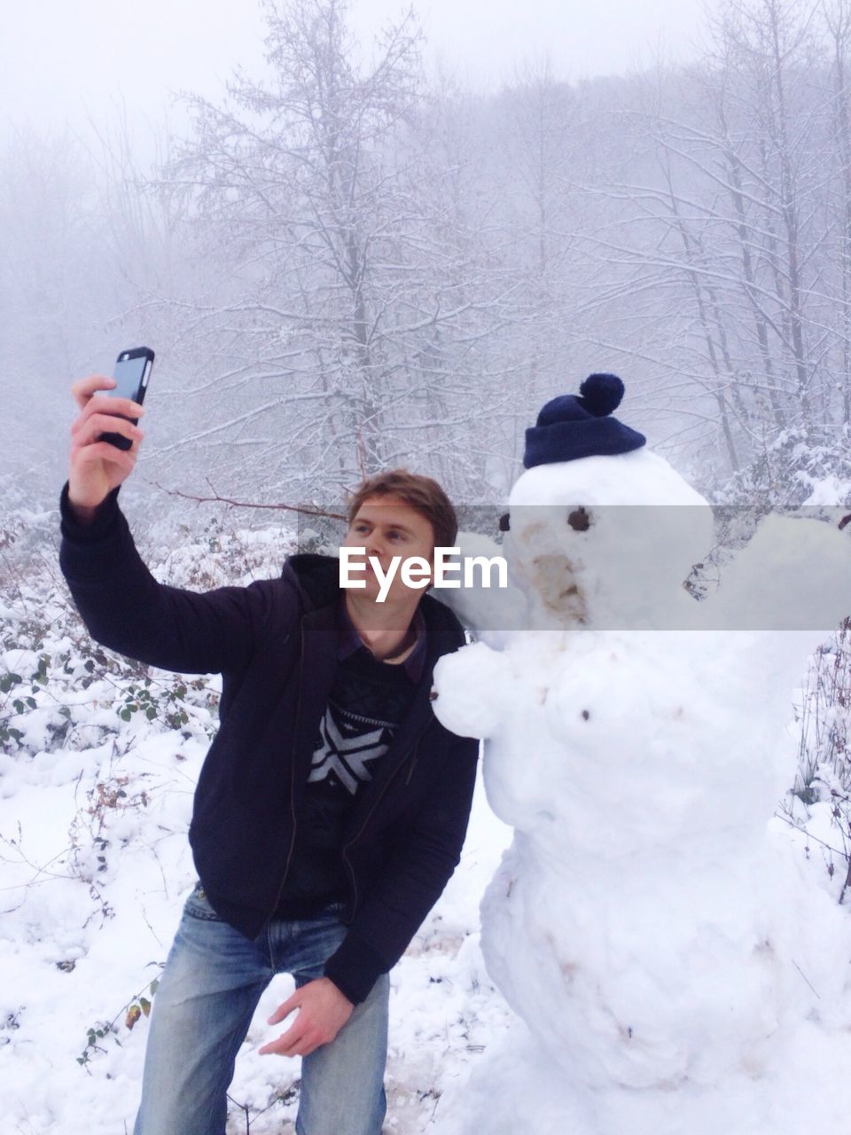 Man clicking selfie with snowman through mobile phone on field