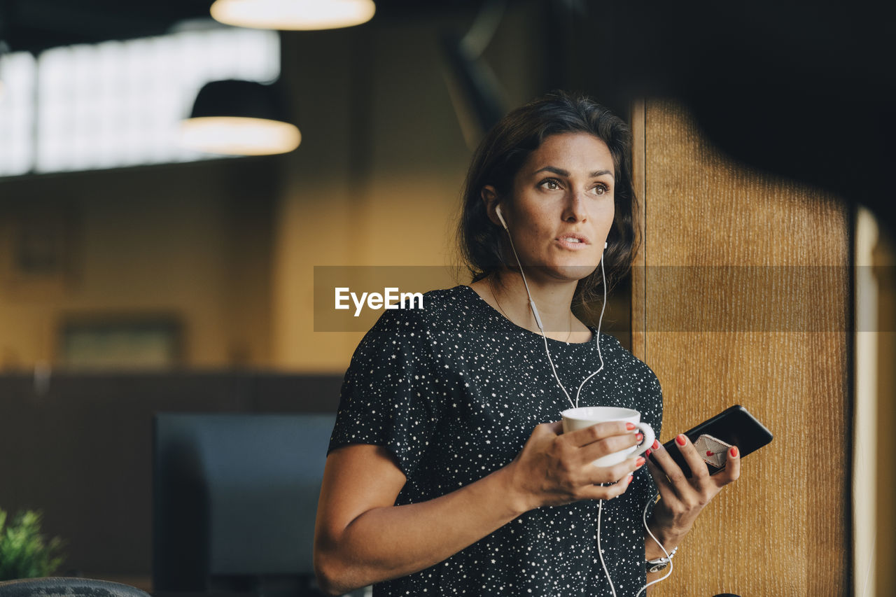 Female executive holding coffee cup while talking with earphones during phone call in office