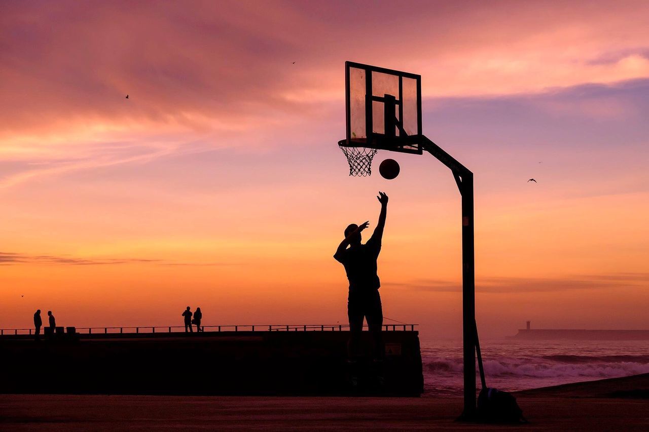 Silhouette of person playing basketball and people at seaside against sky during sunset