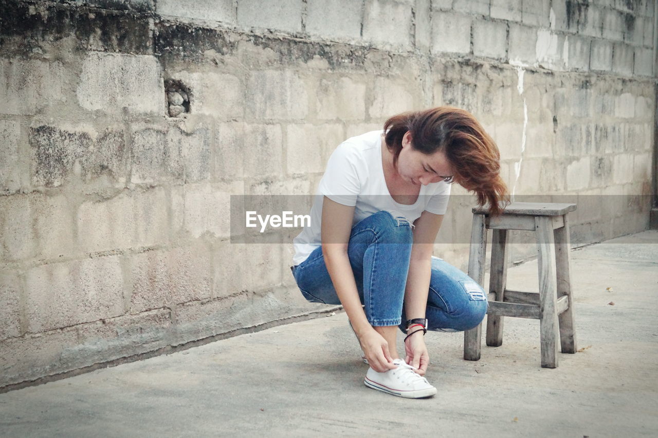 Young woman tying shoes while crouching against wall