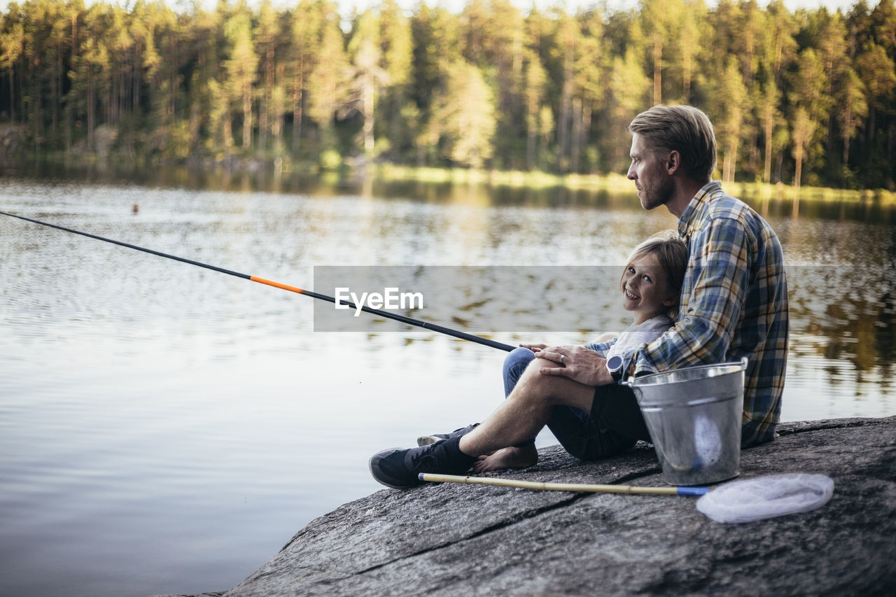 Portrait of smiling daughter fishing with father while sitting by lake