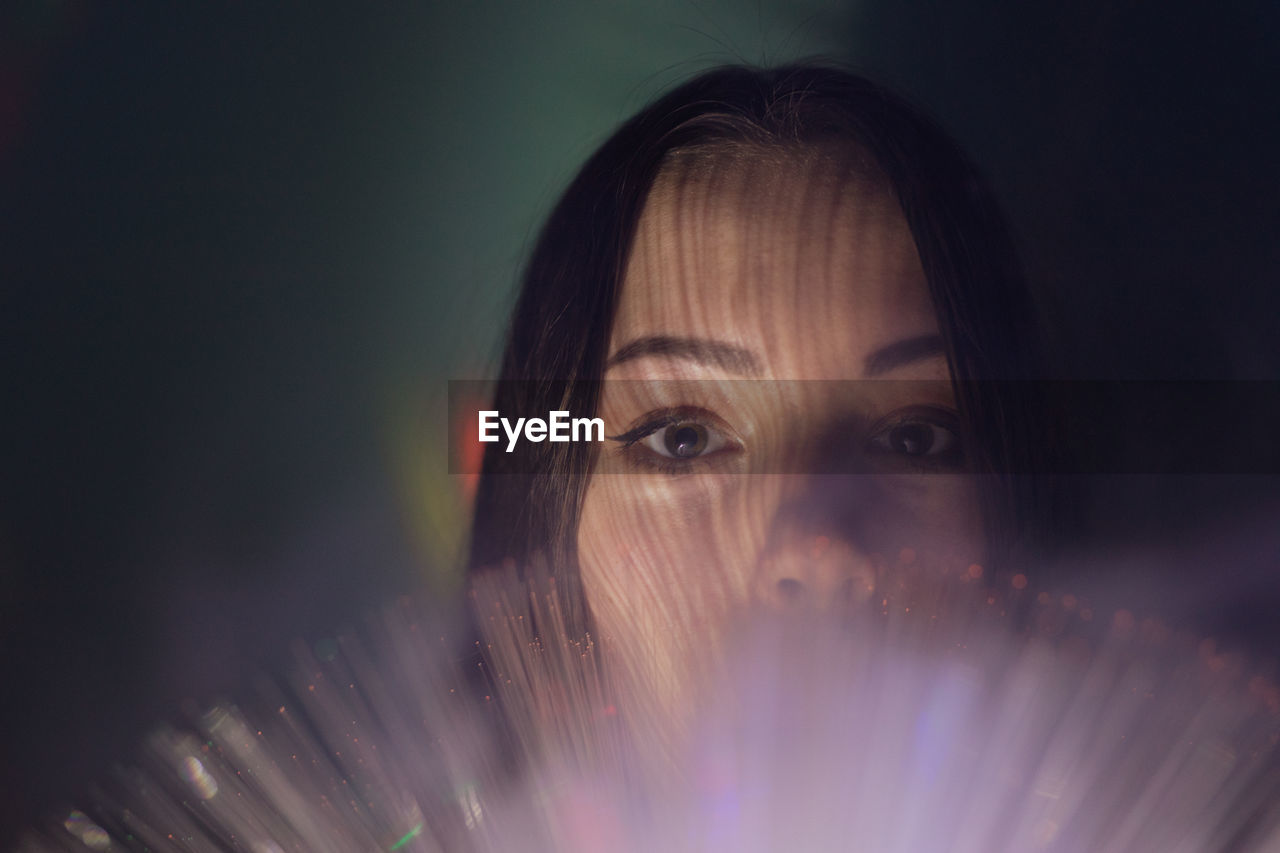 Close-up portrait of young woman holding illuminated fiber optic in darkroom