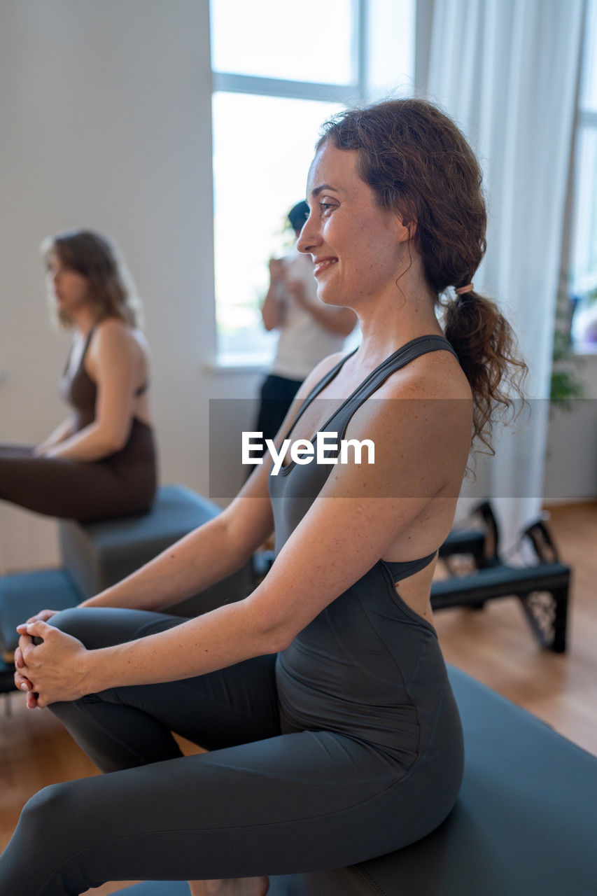 women, adult, exercising, lifestyles, indoors, wellbeing, physical fitness, sports clothing, sports, sitting, female, yoga, side view, gym, young adult, stretching, clothing, arm, health club, human leg, relaxation, flexibility, focus on foreground, two people, person, limb, smiling, togetherness, yoga studio, body care, brown hair, sports training, leisure activity, strength, looking, three quarter length, full length, mature adult, practicing, tank top, vitality, ponytail, exercise mat, hairstyle, balance, body conscious, happiness, concentration, room, healthcare and medicine, pilates, posture