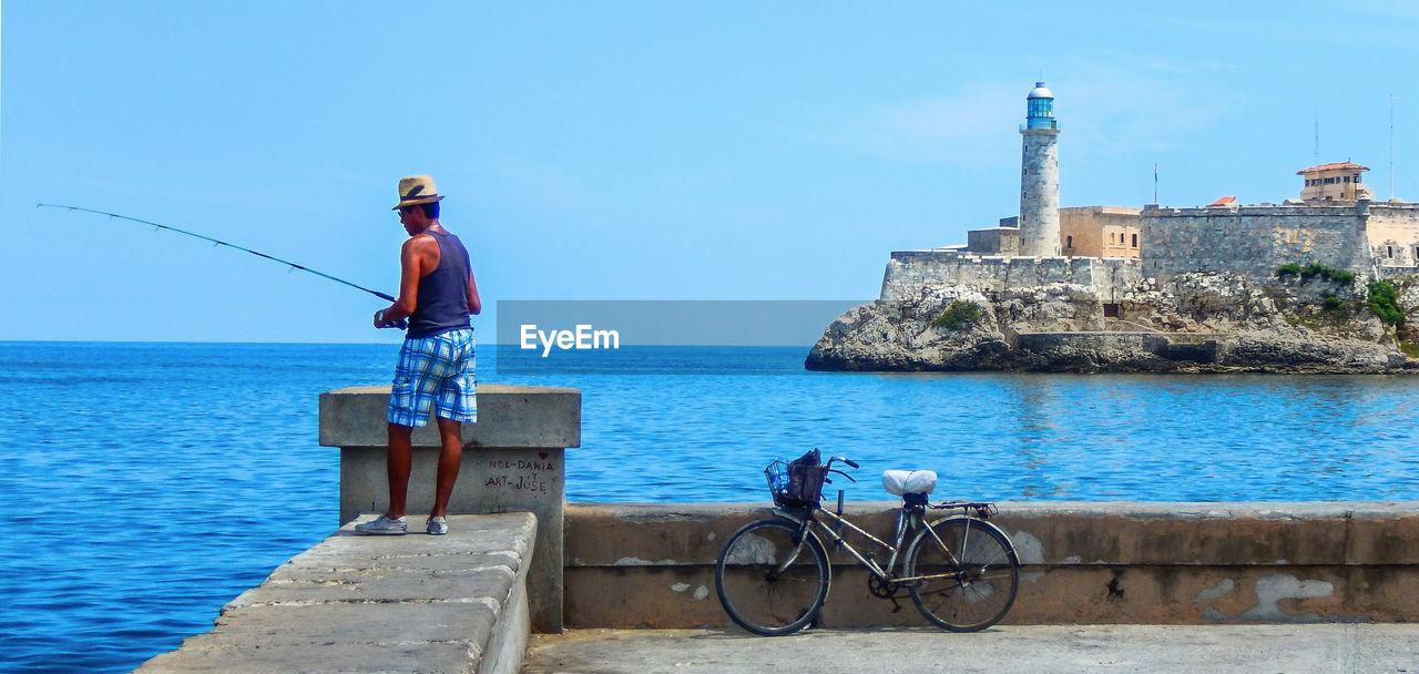 MAN WITH BICYCLE STANDING BY SEA AGAINST SKY