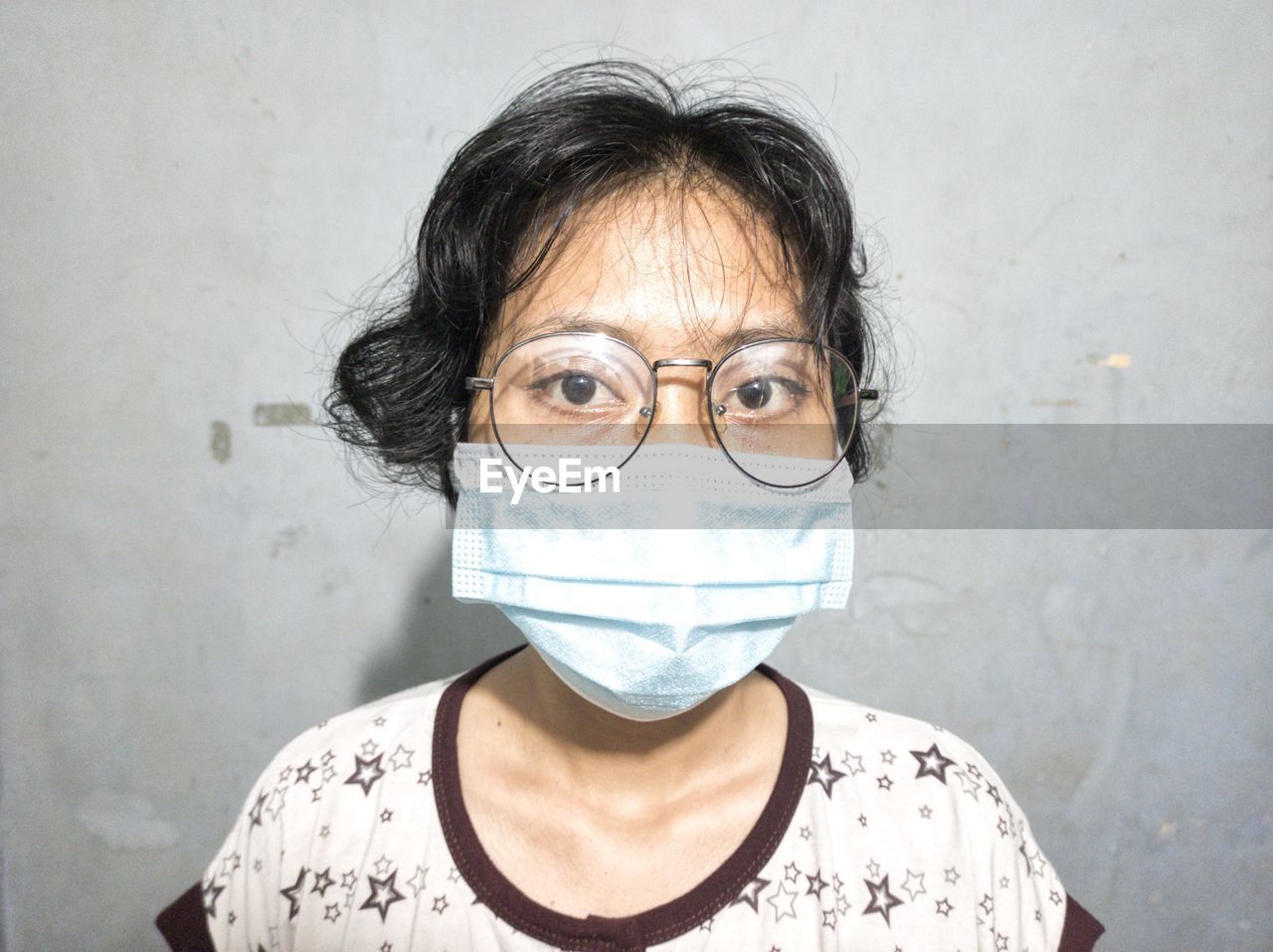 Portrait of woman wearing surgical mask and eyeglasses leaning against a wall