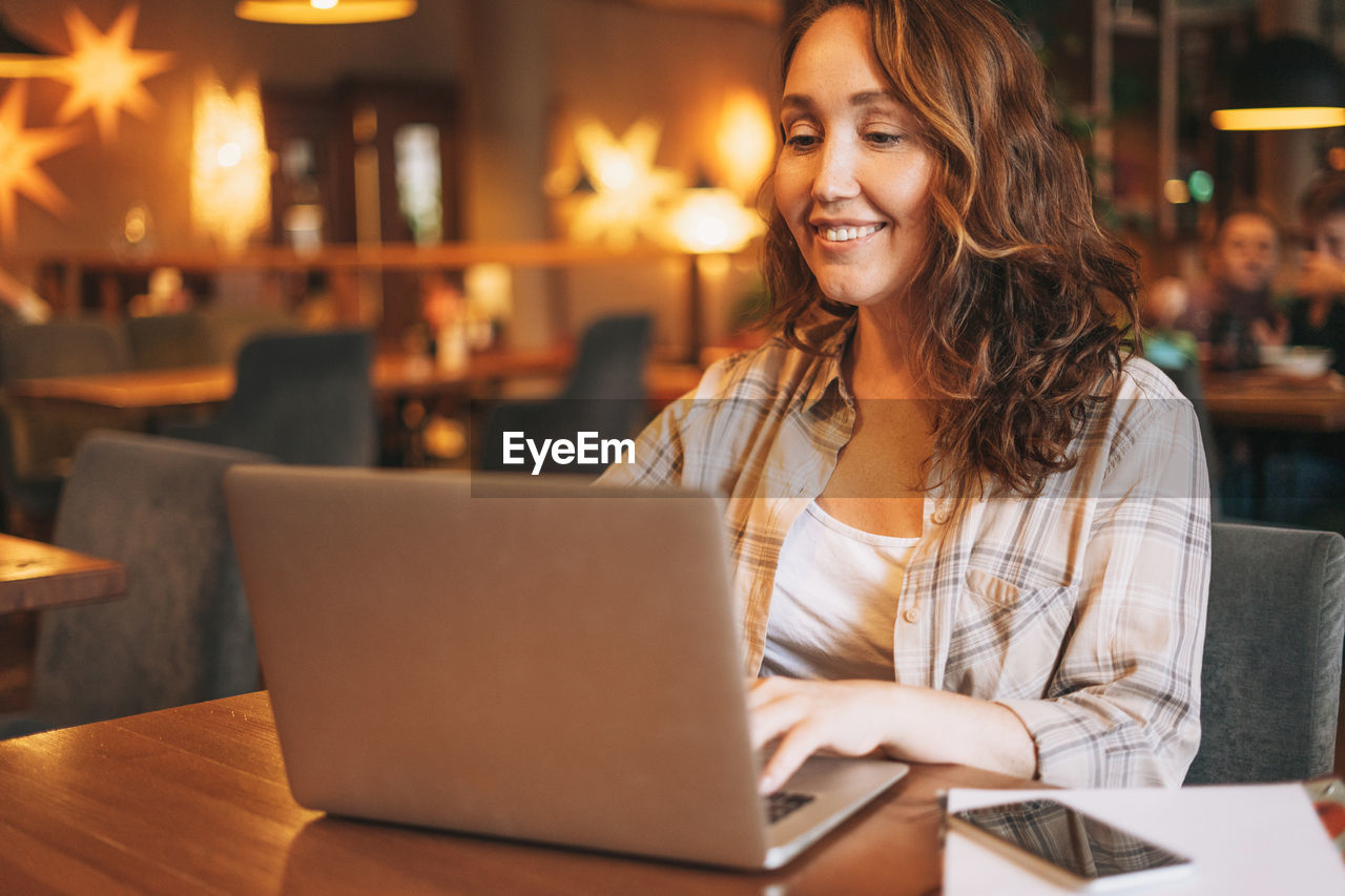 Adult charming brunette woman in plaid shirt working with laptop at cafe