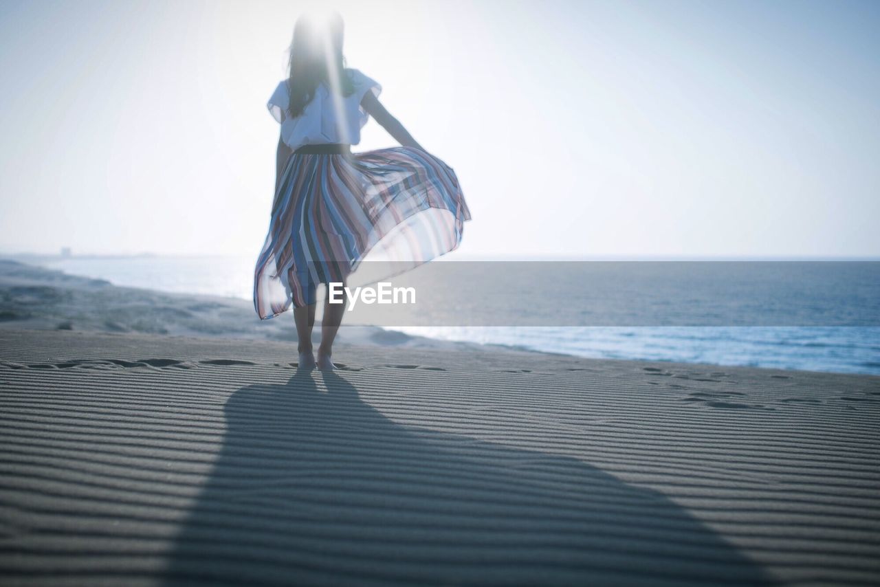 Woman in dress standing on shore at beach against sky during sunny day