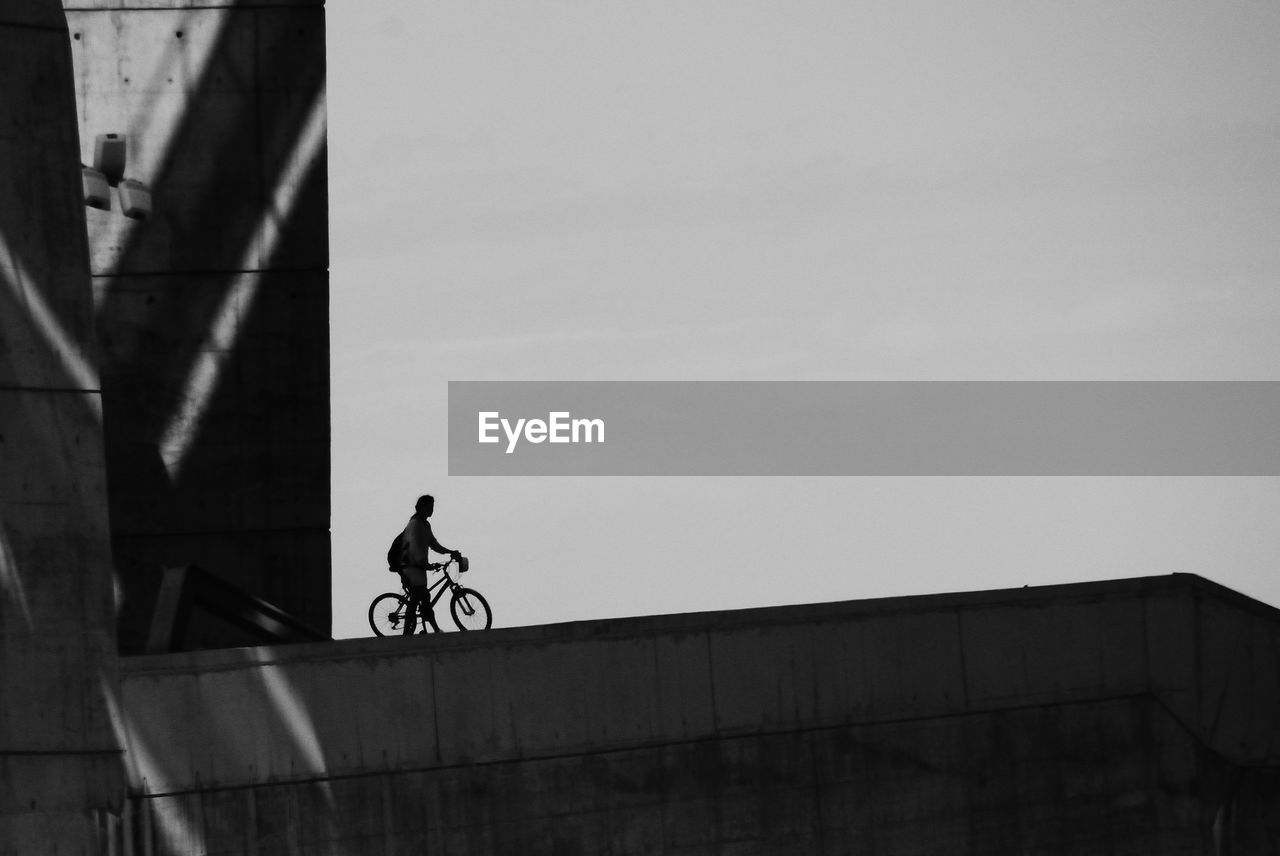 MAN RIDING BICYCLE ON BRIDGE AGAINST SKY