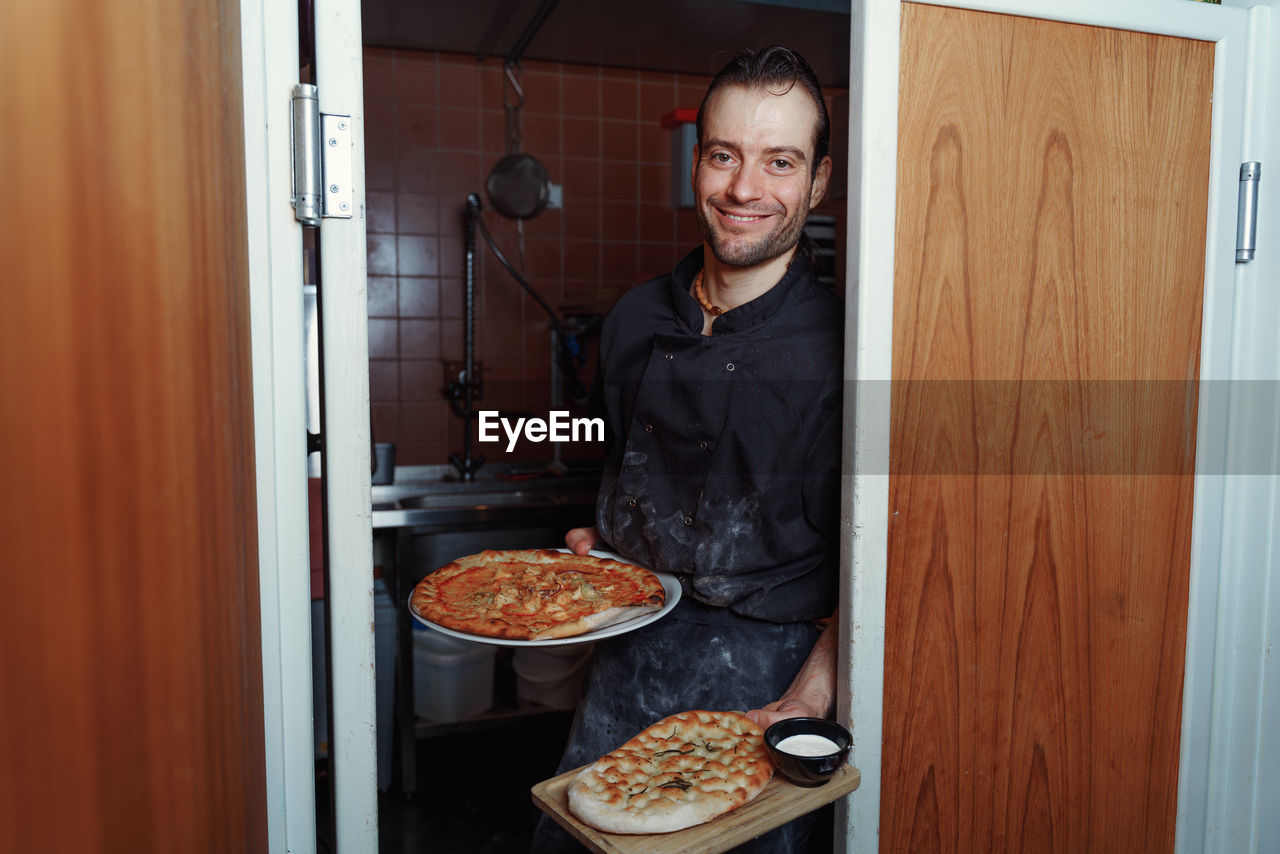 Smiling male chef looking at camera and showing a pizzas against kitchen 