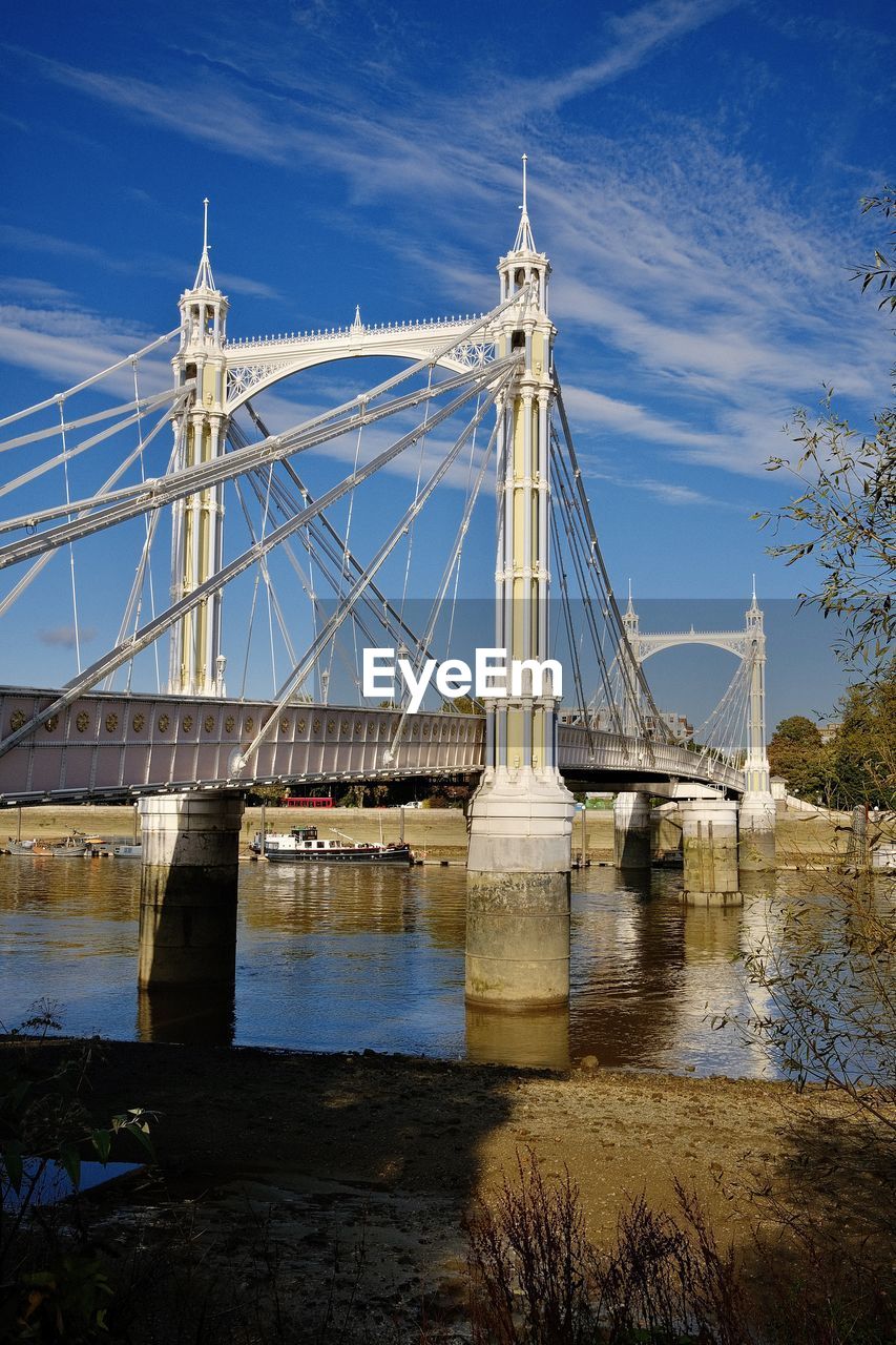 View of albert bridge from battery park, london. suspension bridge over river against cloudy sky.