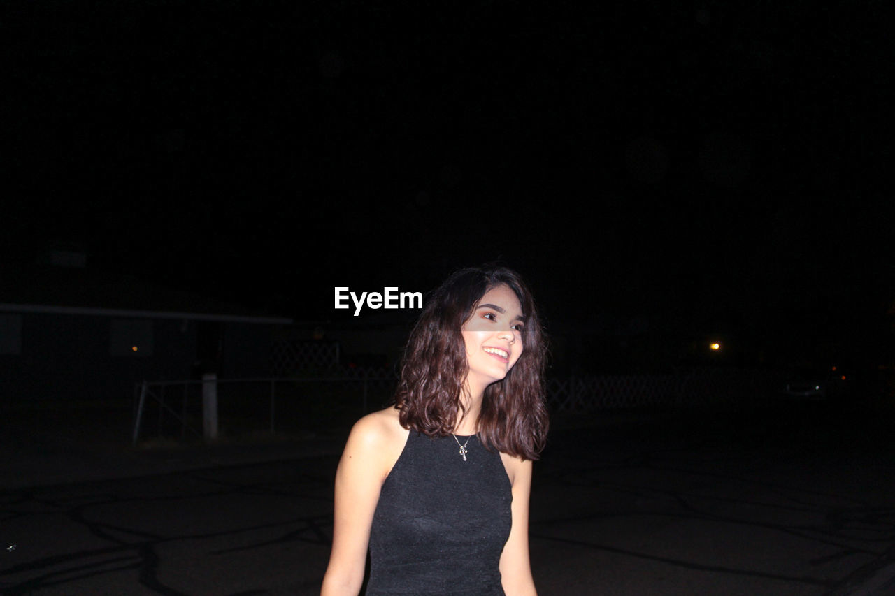 Smiling beautiful woman looking away while standing against sky at night