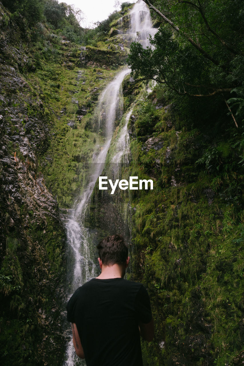 REAR VIEW OF MAN LOOKING AT WATERFALL