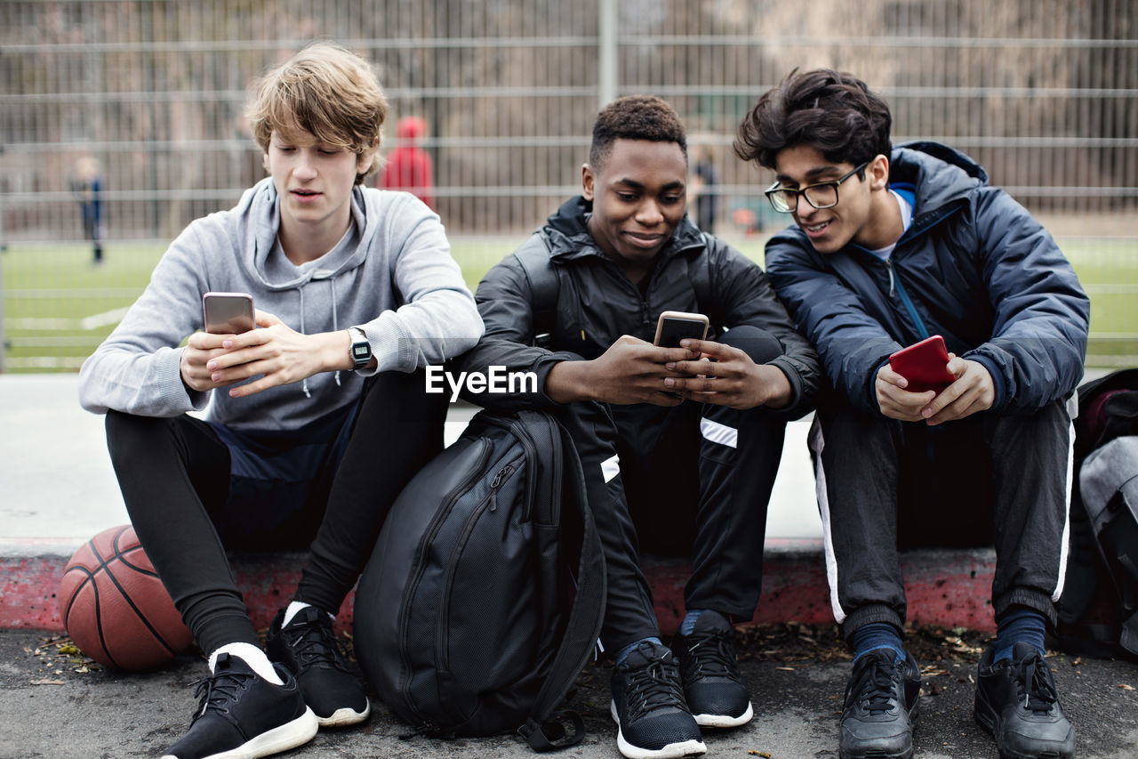 Social media addicted friends using mobile phones while sitting on sidewalk after basketball practice in city