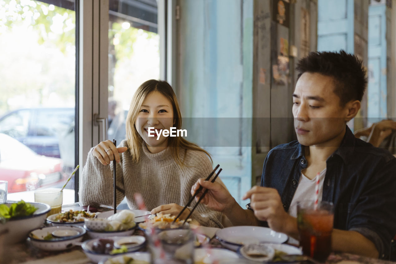 Portrait of smiling woman with chopsticks sitting with male friend having food at restaurant