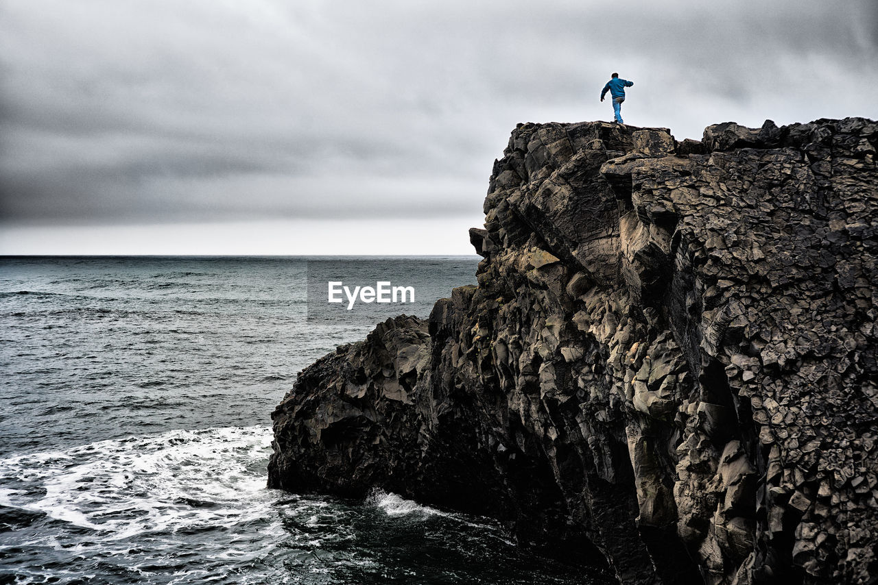 Rear view of man walking on cliff by sea against cloudy sky