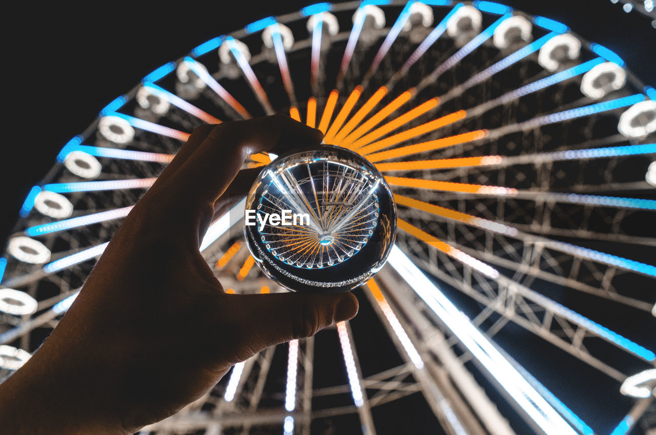 Cropped hand holding crystal ball against illuminated ferris wheel at night