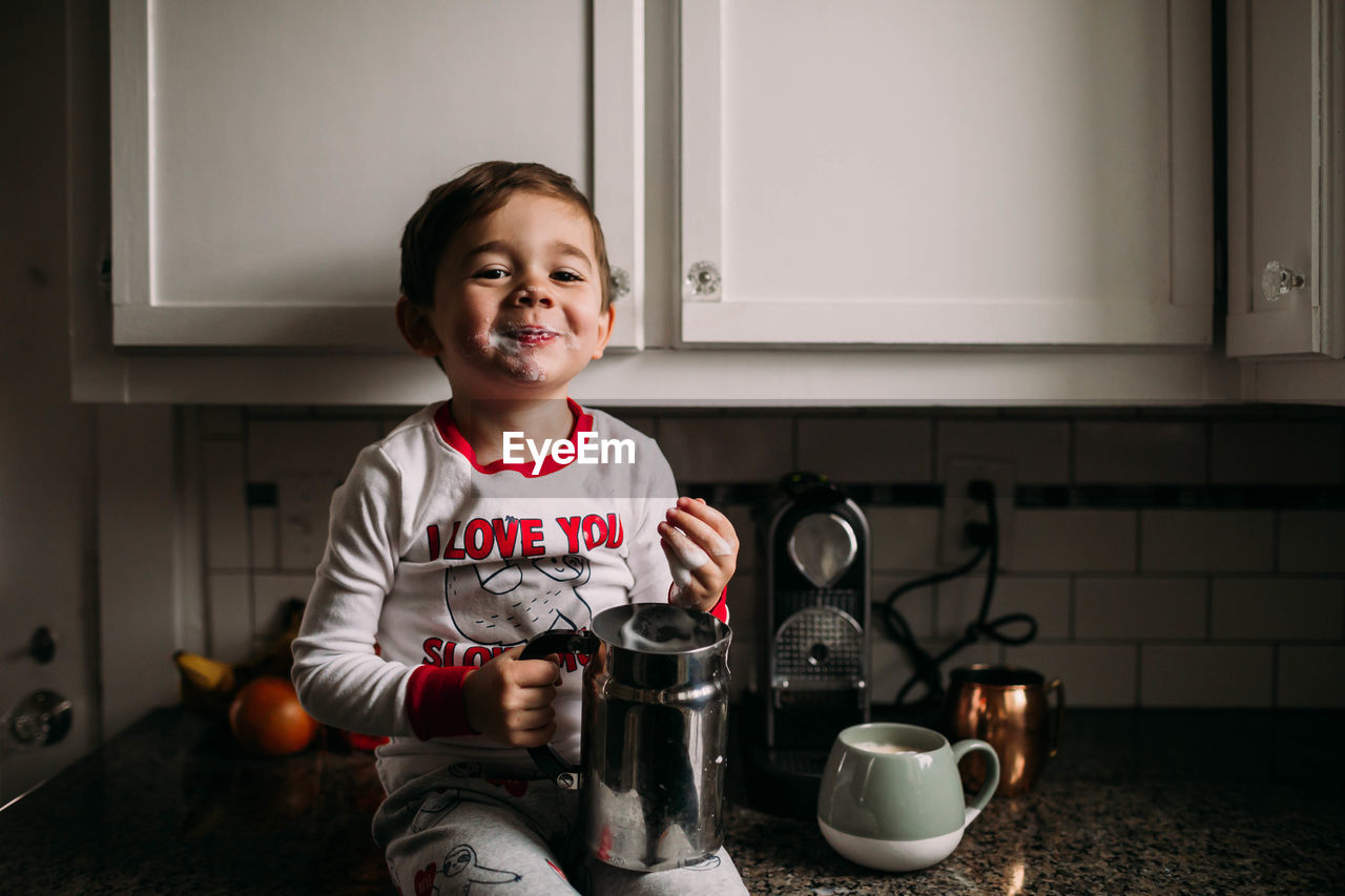 Young boy sitting on kitchen counter with frothed milk on face