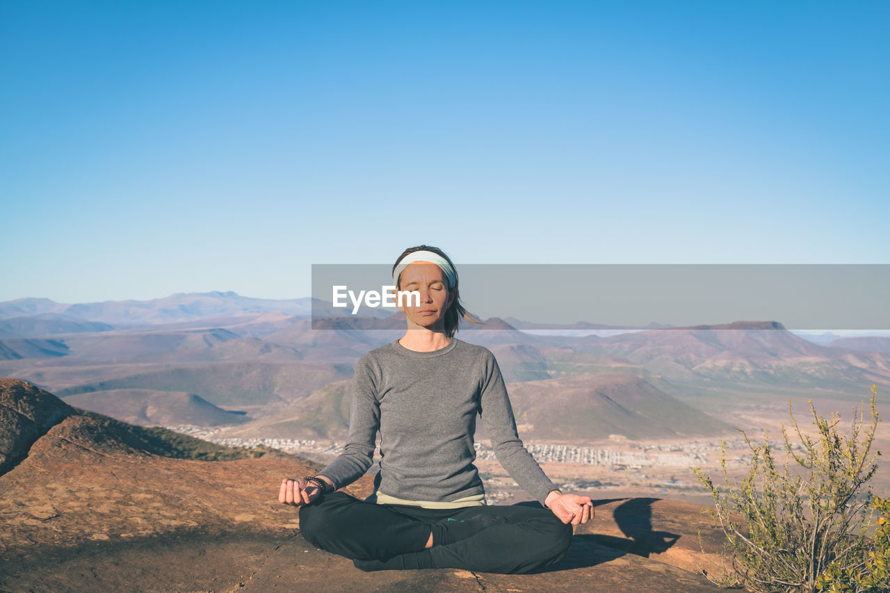 Woman practicing yoga on rock at mountain peak against blue sky during sunny day