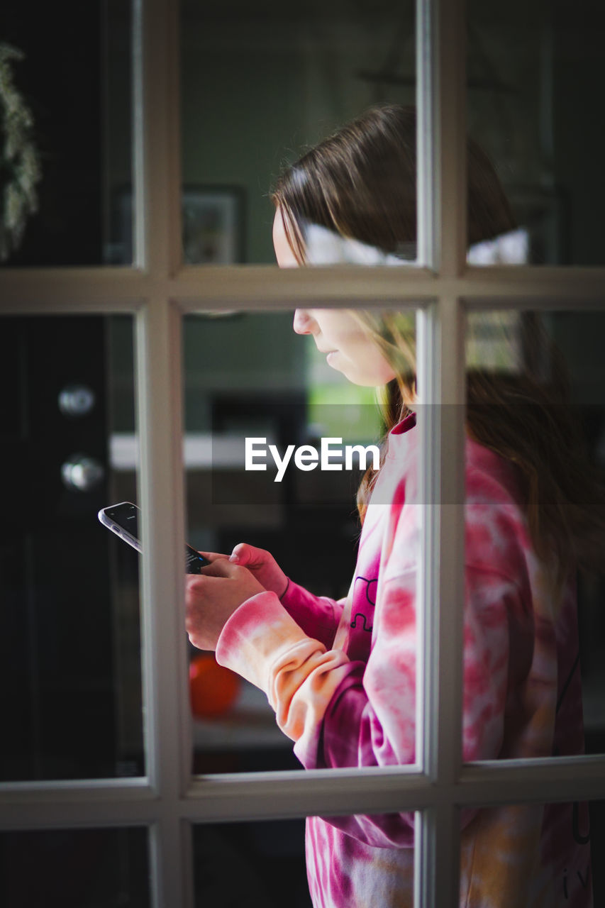 Side view of girl using smart phone seen through window