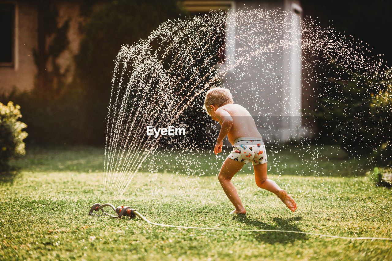 Young white boy running under the water from the sprinkler in garden