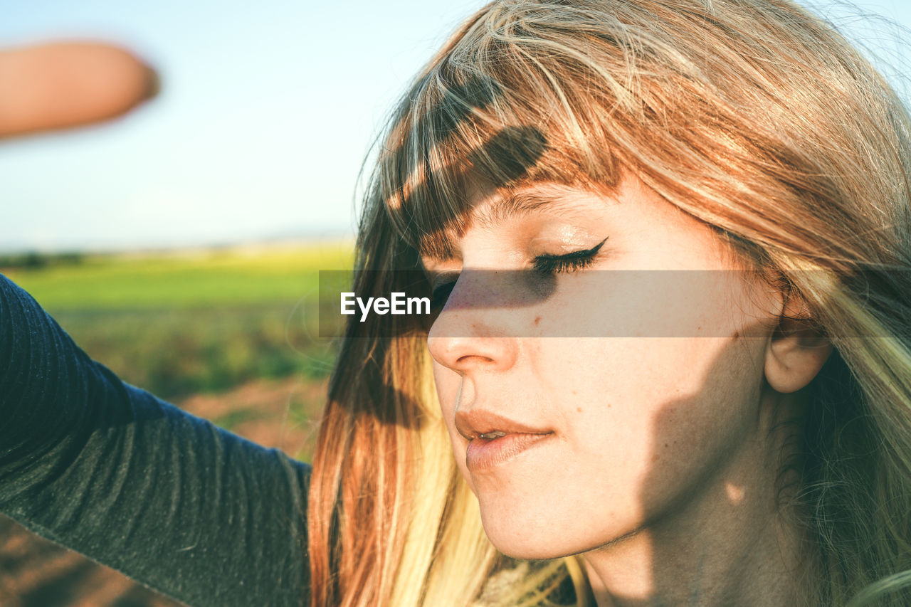 Close-up of young woman shielding eyes while standing at field against sky on sunny day