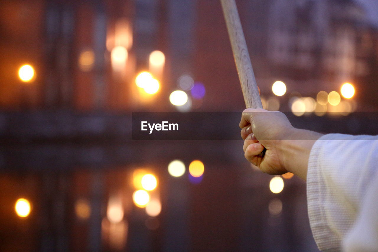 Cropped image of man practicing martial arts with sword against illuminated canal in city during sunset