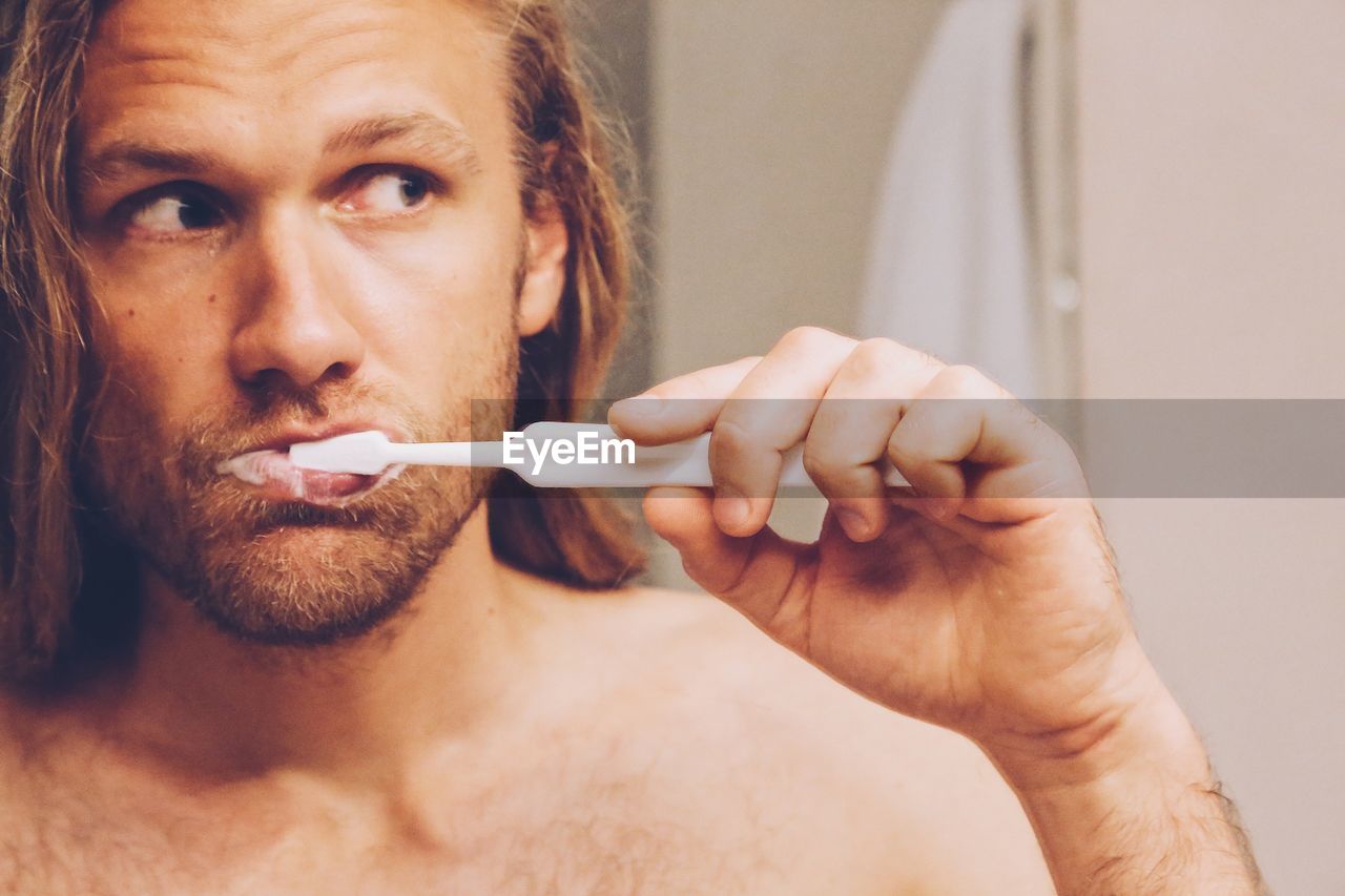 Close-up of shirtless man looking away while brushing teeth in bathroom