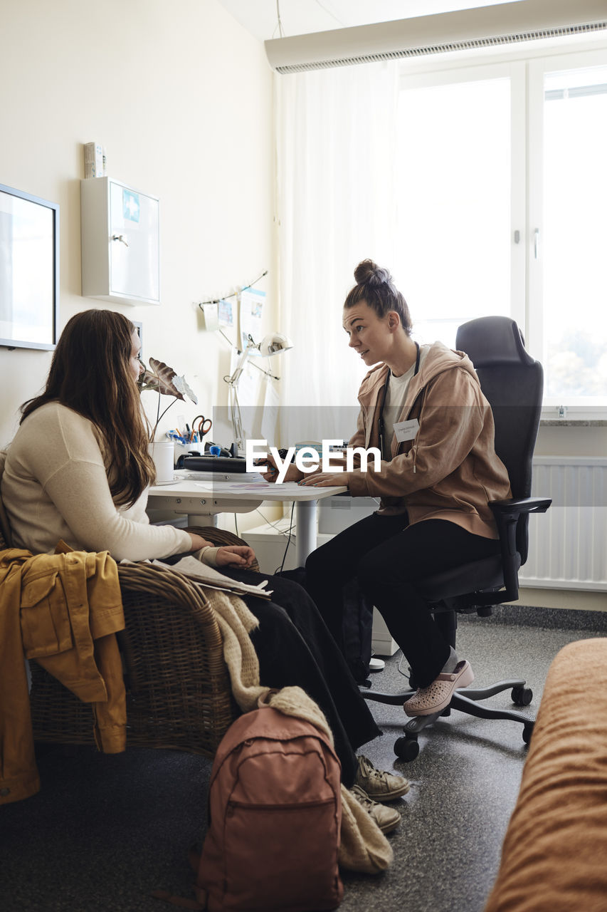 Full length of non-binary school nurse discussing with female student in office