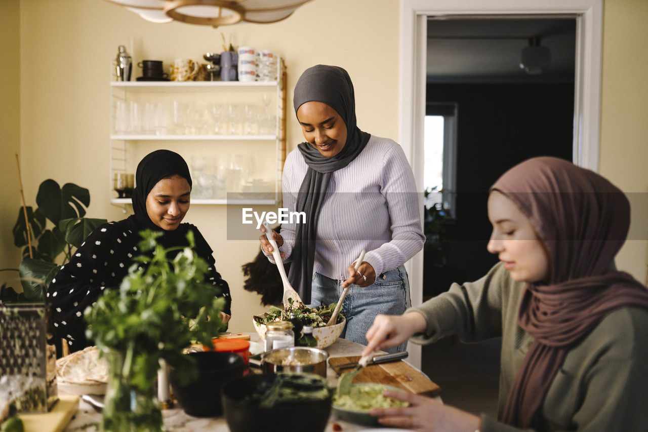 Smiling woman making salad while standing by friends preparing food at table in kitchen