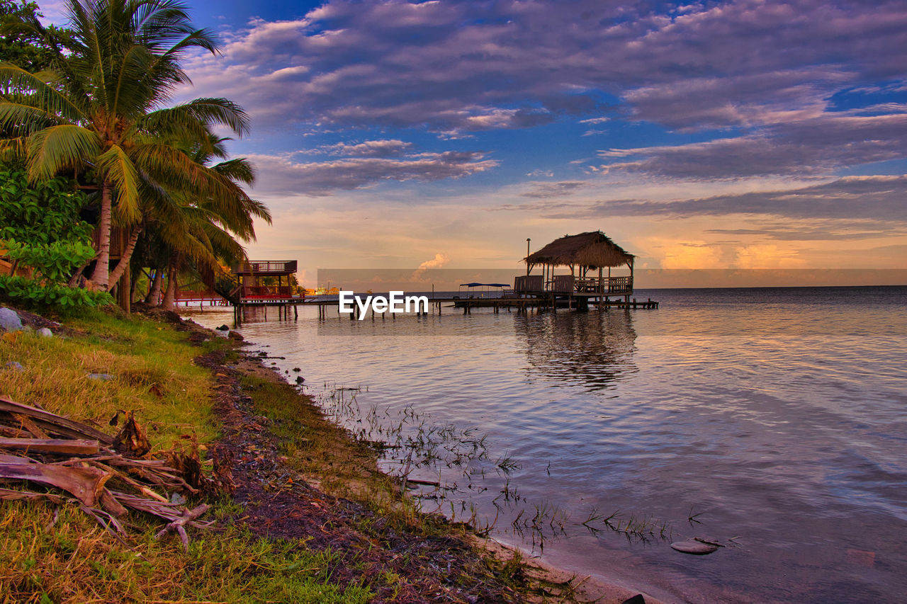 water, sky, sea, architecture, sunset, shore, tropical climate, beach, nature, cloud, palm tree, built structure, coast, land, beauty in nature, travel destinations, dusk, scenics - nature, reflection, evening, horizon, ocean, travel, tranquility, tree, vacation, trip, environment, body of water, holiday, plant, building, building exterior, landscape, tourism, hut, tranquil scene, house, no people, outdoors, idyllic, bay, seascape, coconut palm tree, coastline, stilt house, horizon over water, blue, sunlight, pier, twilight, tropics, island