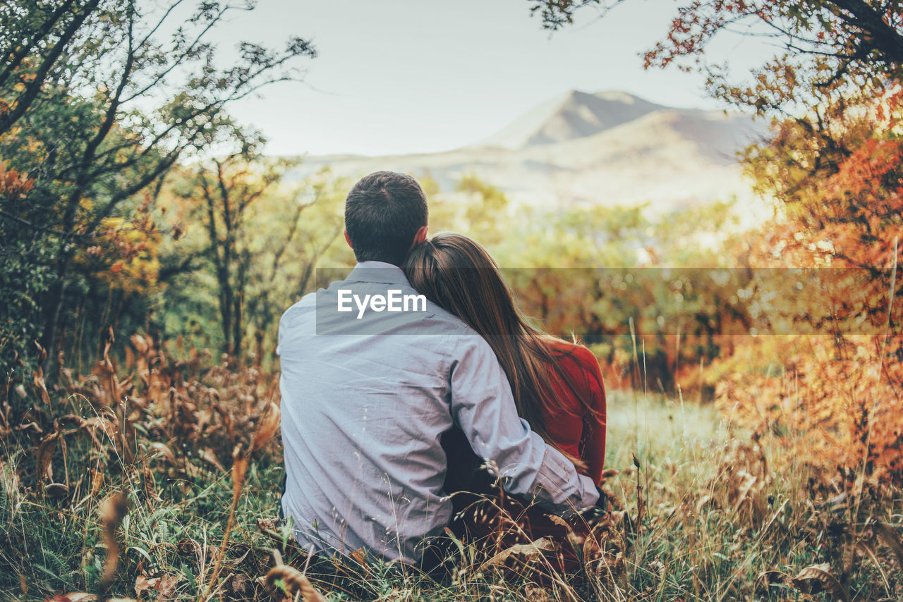 REAR VIEW OF COUPLE SITTING ON LAND BY PLANTS