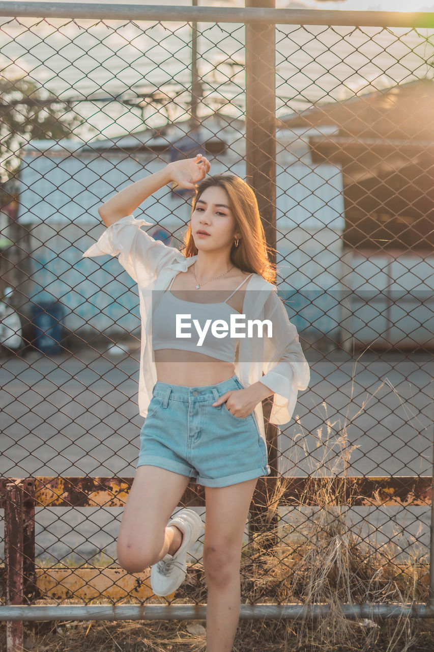 Full length of young woman standing by chainlink fence